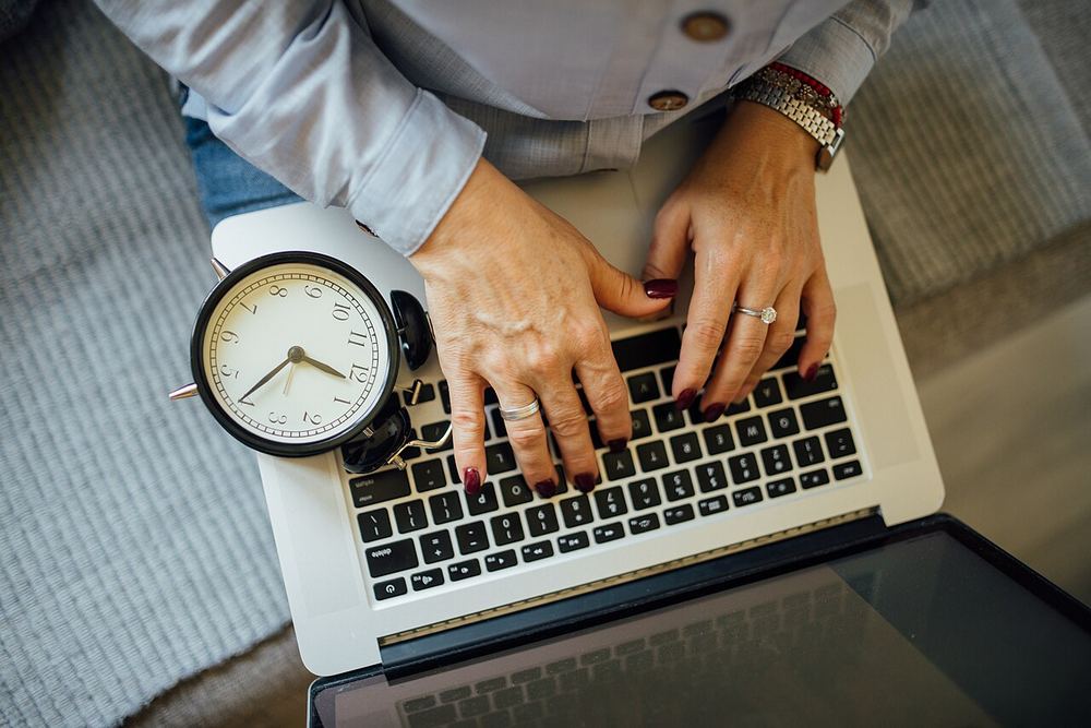 Manos escribiendo en un portátil junto a un reloj, esperando